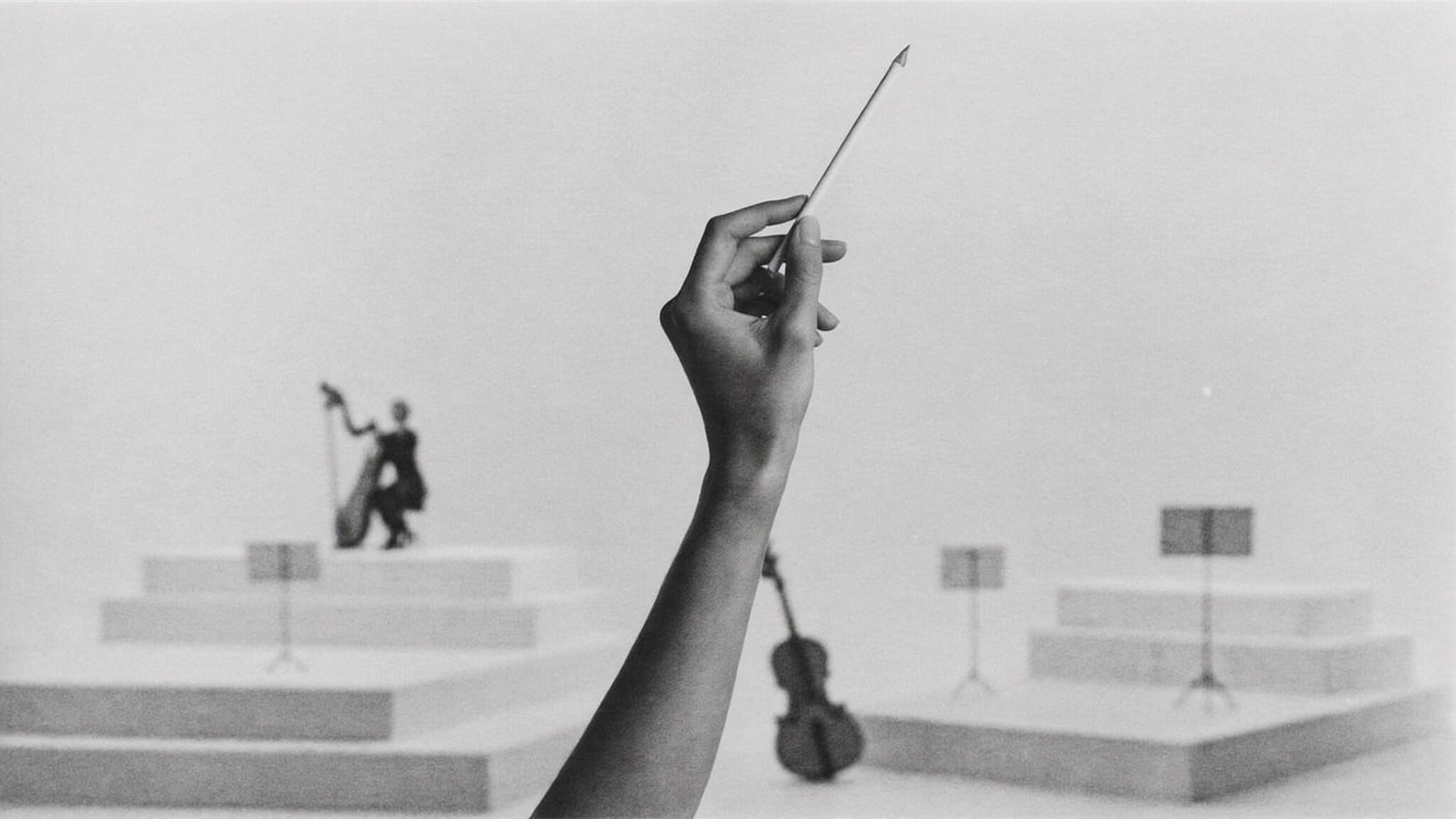 A hand holding a pencil raised like a conductor's baton, before a small orchestra of instruments